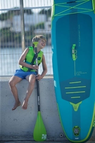 Un copil care râde, poartă o vestă de salvare și un costum de baie, cu o placă în mână, stă pe un perete pe care se sprijină un paddleboard gonflabil SUP.