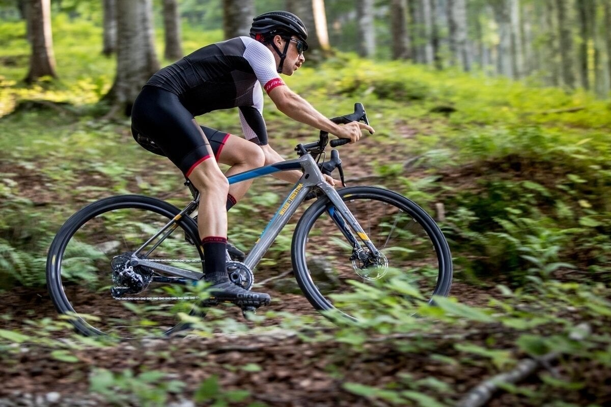 Bici da gravel Wilier Jena con telaio in carbonio scuro, freno a disco idraulico e manubrio sportivo con un ciclista che va in foresta.