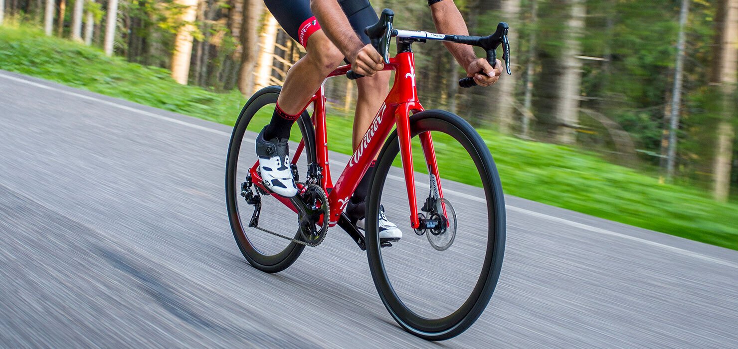 Cestný bicykel Wilier s ľahkým červeným karbónovým rámom, športovými riadidlami - baranmi so sediacim pretekárom pohybujúci sa vo vysokej rýchlosti po asfaltovej ceste.