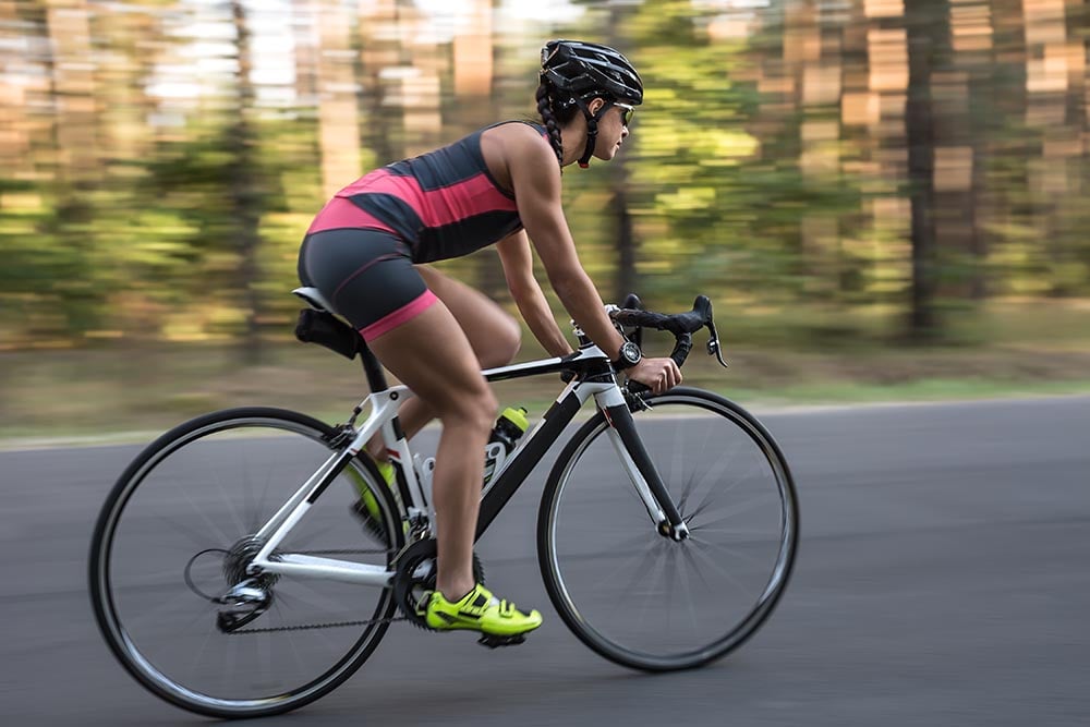 Egy fiatal lány kerékpáros ruhát és biciklis sisakot viselve nagy sebességgel halad egy országúti kerékpárral az aszfaltúton.