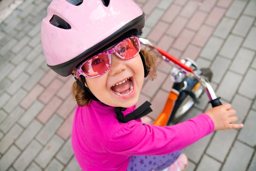 Dievčatko s veselým výrazom vo veku 4 roky s ružovou cyklistickou prilbou, ružovými slnečnými okuliarmi v ružovom oblečení jazdí po chodníku na detskom bicykli.